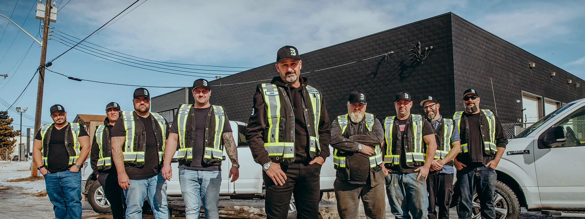 A diverse group of professional male construction or field workers standing together in front of a commercial building and work trucks, wearing matching high-visibility safety vests and branded hats.